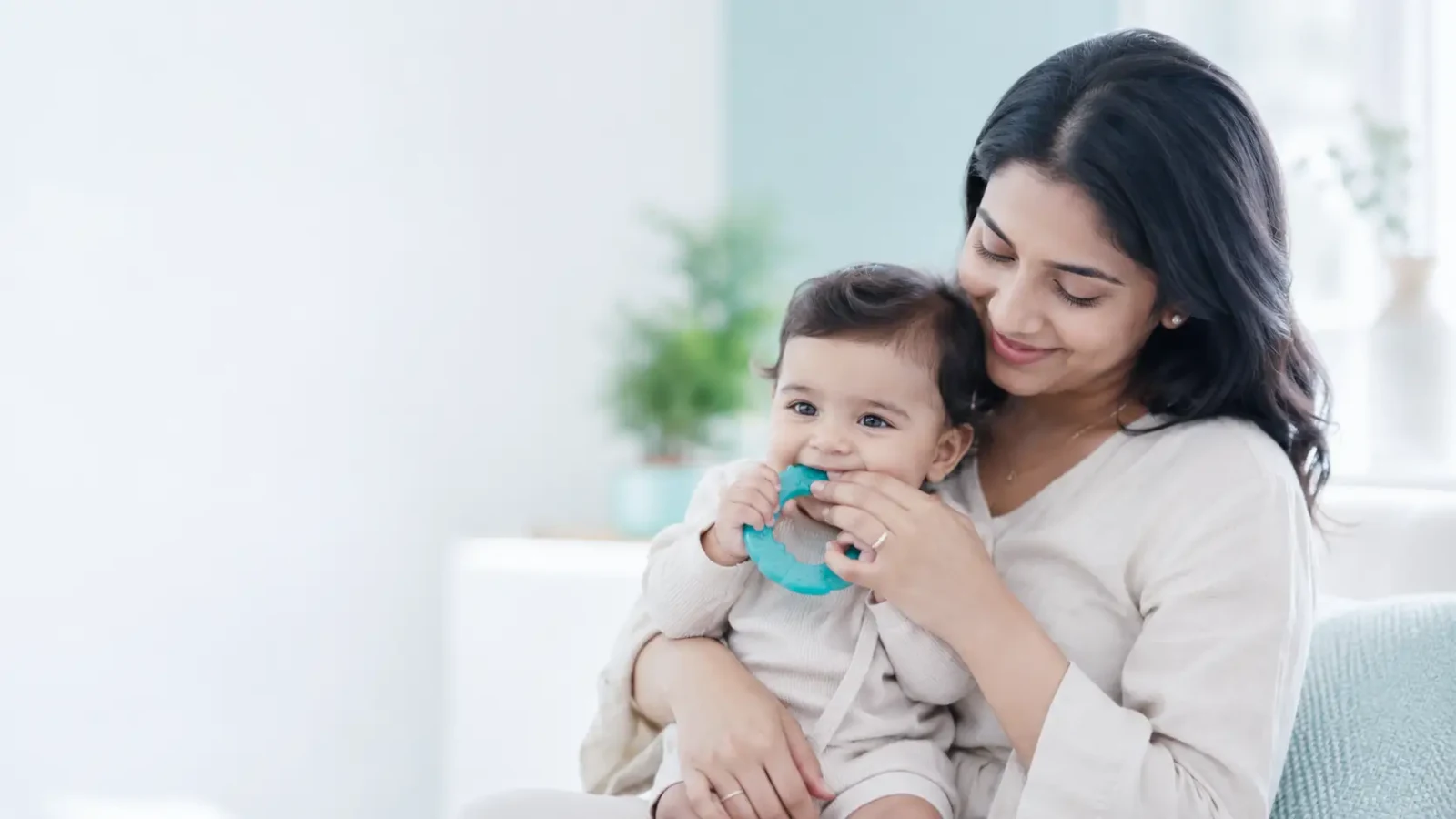 A mother holding her smiling baby, who is chewing a pale teal silicone teething ring.