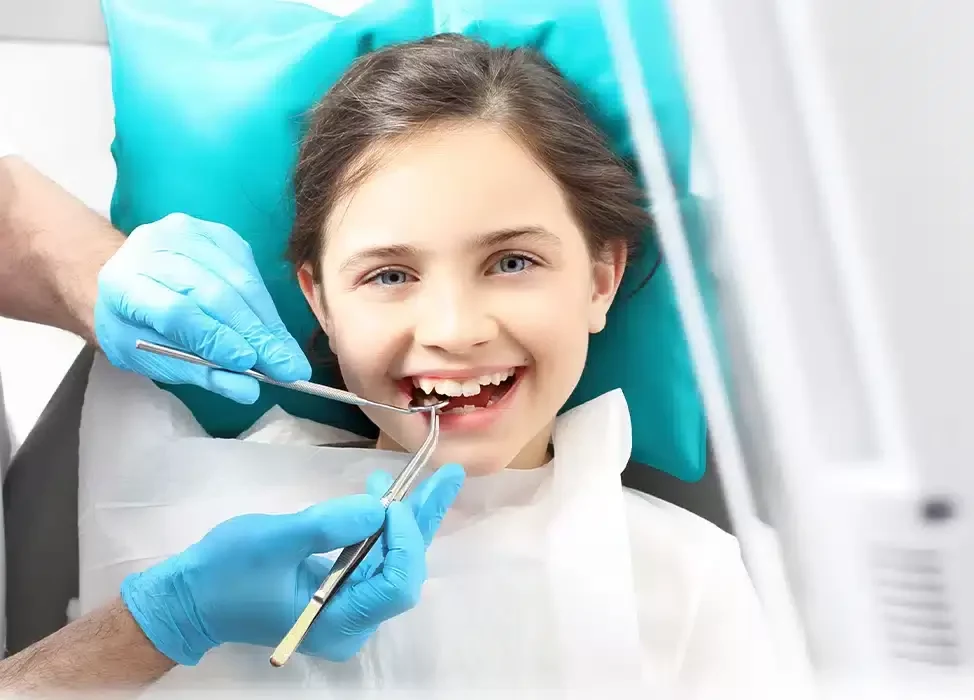 A child smiling in a dental chair while a dentist in blue gloves examines her teeth with a mirror and probe.