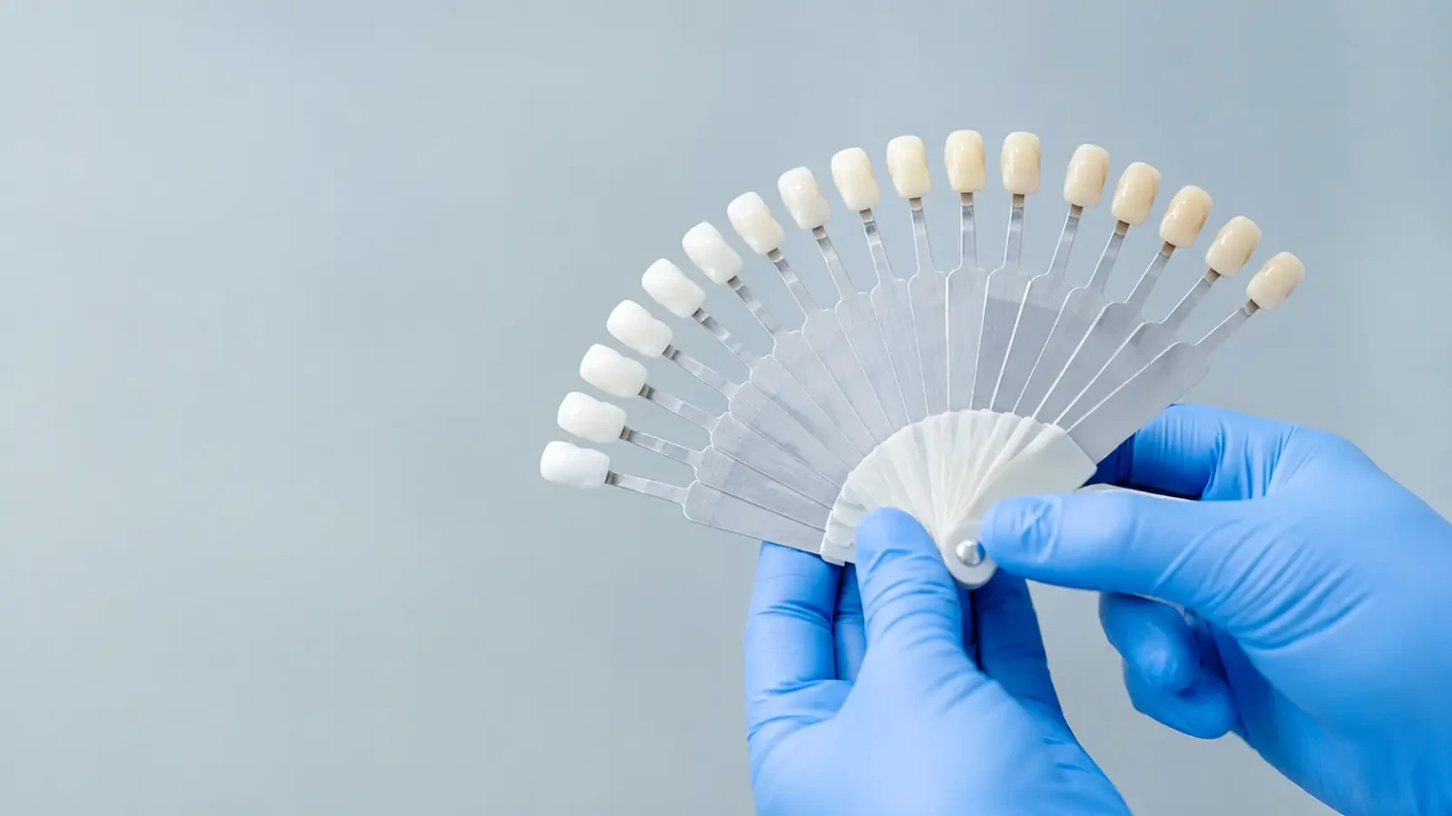 A dentist in blue gloves holding a fanned composite shade guide showing tooth-coloured shade tabs.