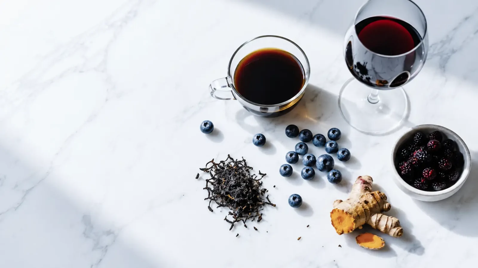 Black coffee, red wine, blueberries, loose tea, turmeric and dark berries on a white marble surface.
