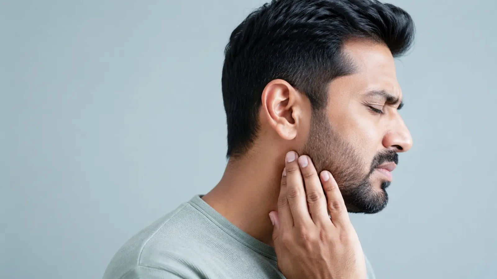 A man in profile with his eyes closed, pressing his fingers against his jaw just below the ear.
